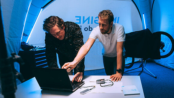 One person shows something on the laptop to a second person in the Pit Stop tent